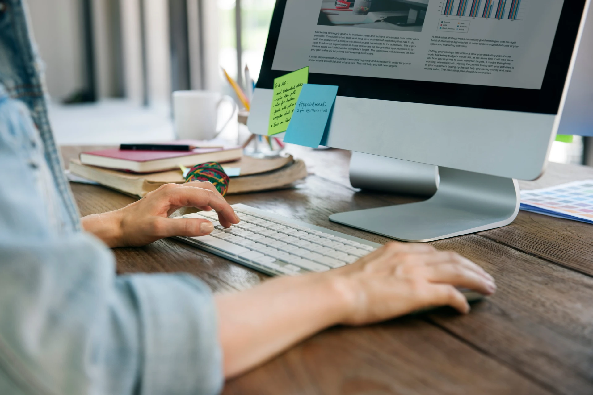 A person seated at a desk with dual monitors displaying text and coding, surrounded by tech equipment and a plant.